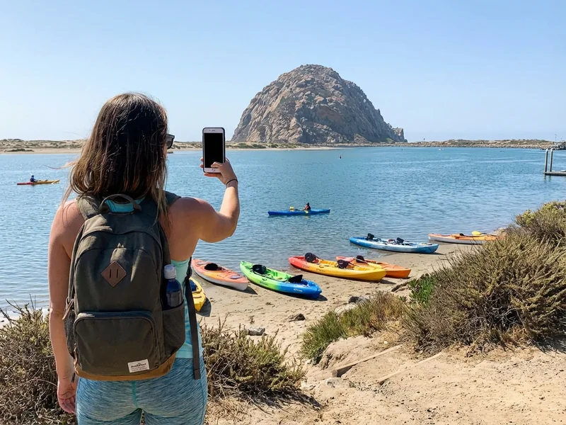 Person holding smartphone with kayak rental search results visible, Morro Rock in background